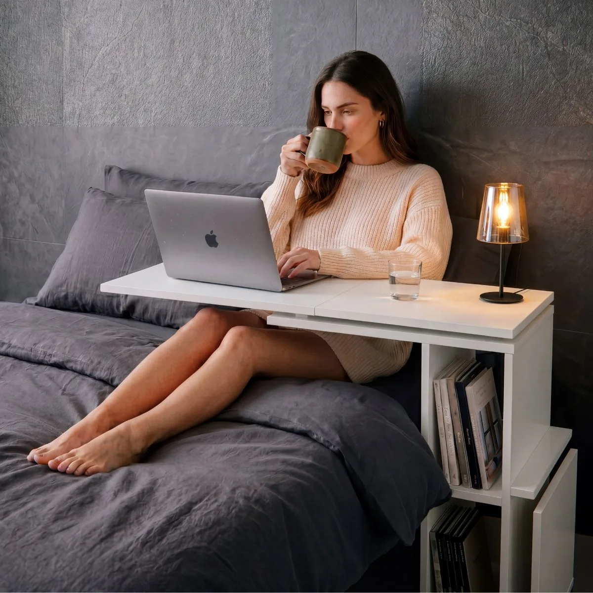 Mujer sentada en la cama tomando un café, con la notebook sobre la bandeja extendida de la mesa de cama blanca; al costado, libros en los estantes